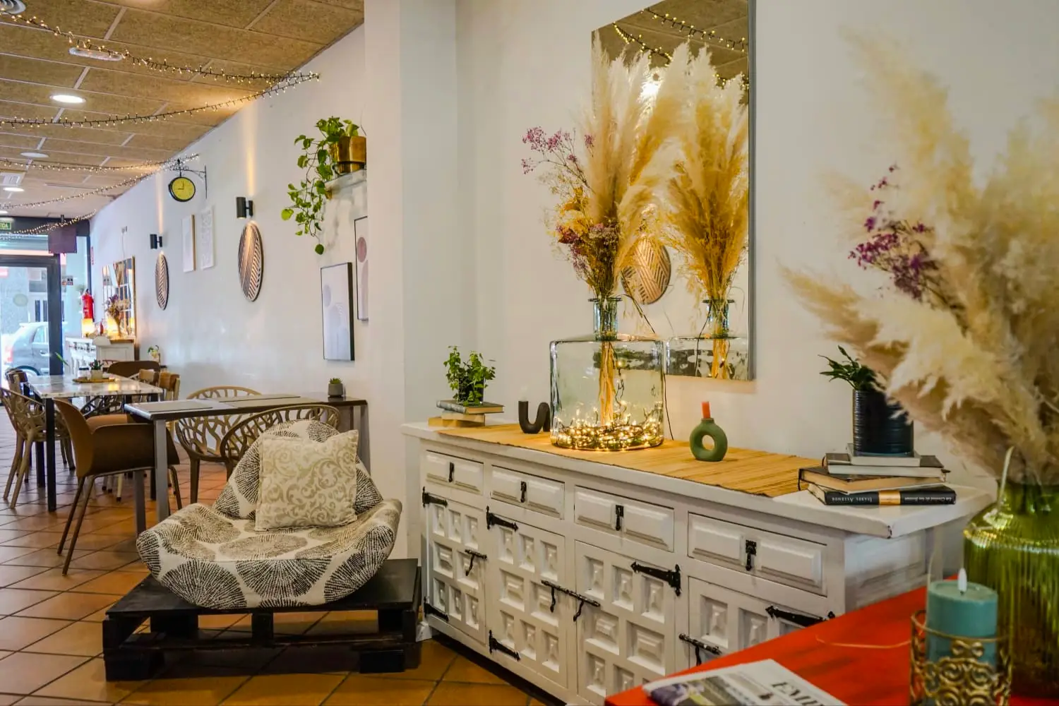 Interior acogedor de La Nonna Brunch en el centro de Figueres, con mesas de madera, sillón bajo y decoración con pampas secas y luces cálidas